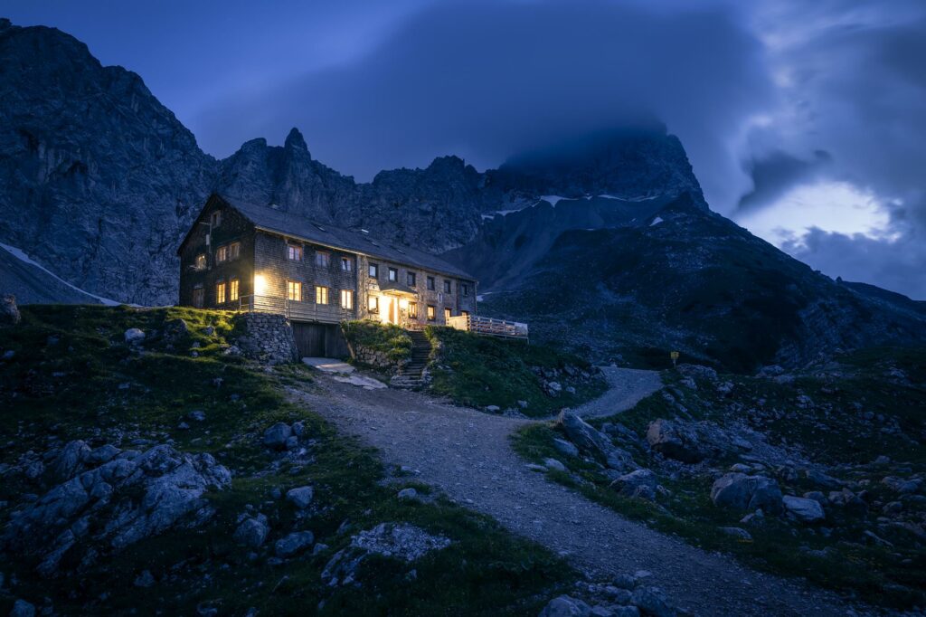 Beispielbild aus dem Magalog "alpinerlebnis", auf der die Berghütte "Lamsenjochhütte" bei angehender Dunkelheit vor steilen Felswänden von innen beleuchtet ist.