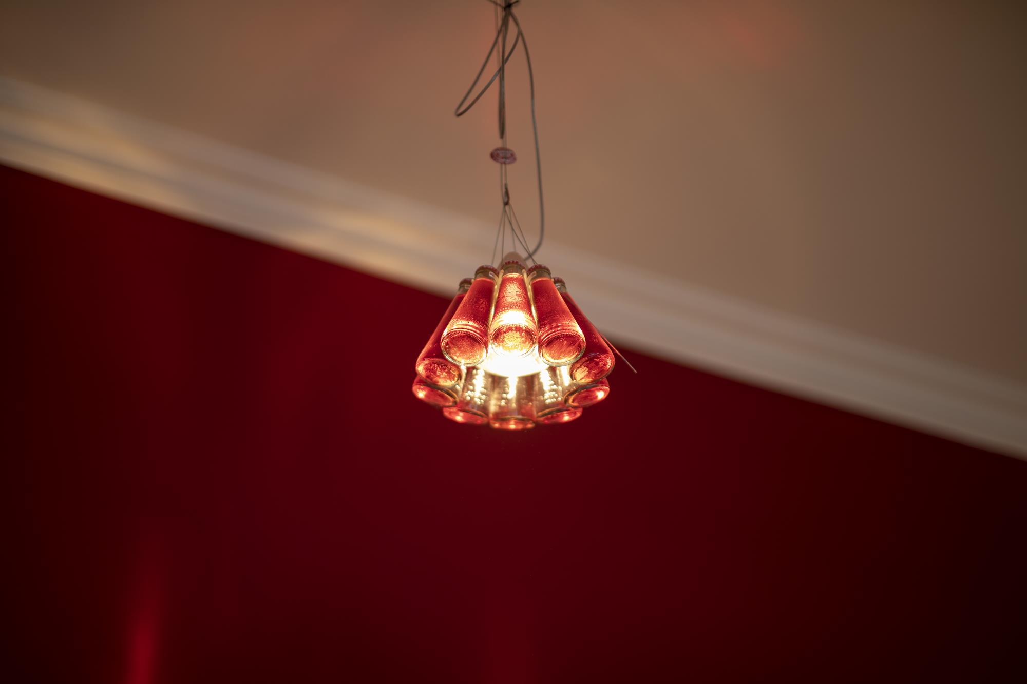Detailaufnahme einer Künstlerlampe, die aus zehn kleinen Sanbitter-Fläschchen besteht. Die Lampe ist an und hängt vor roter Wand und weißer Decke.