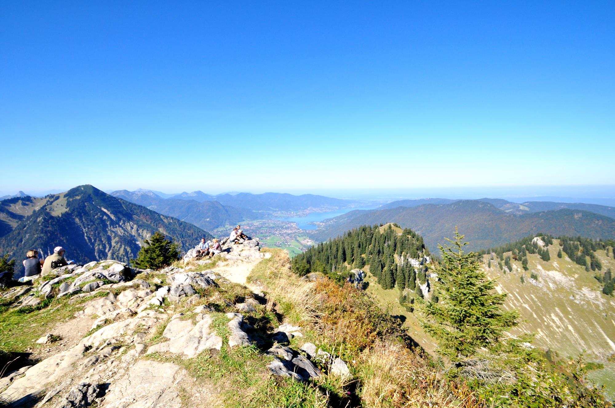 Mehrere Wanderer sitzen bei wolkenlosem Himmel auf einem felsigen Berggipfel. Weit im Hintergrund ist ein blauer See, umrahmt von sanften, grünen Bergen.