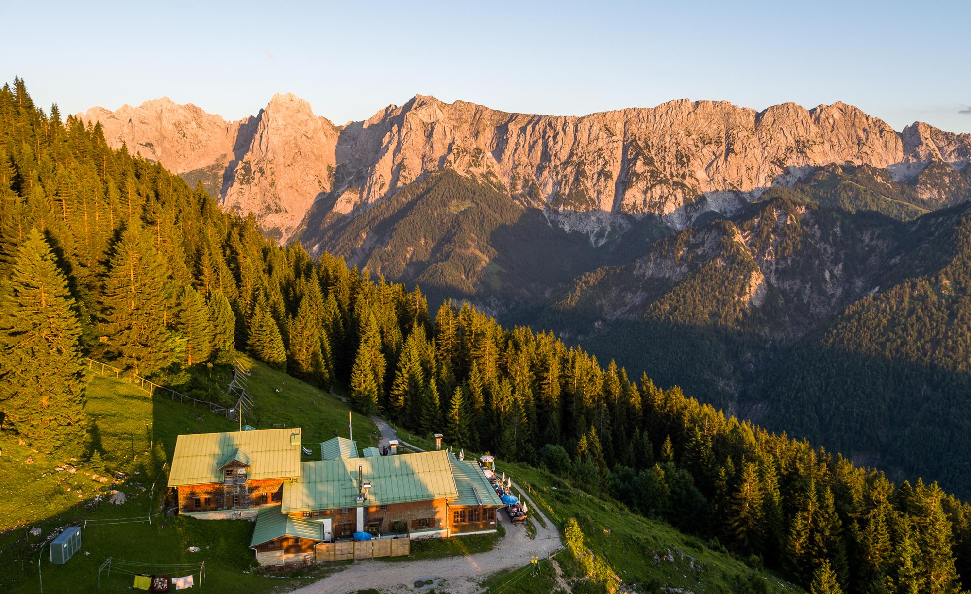 Die Alpenvereinshütte "Vorderkaiserfeldenhütte" bei Sonnenuntergang. Dahinter ein Waldstreifen sowie ein von rötlichem Licht beschienener felsiger Gebirgszug zu sehen.