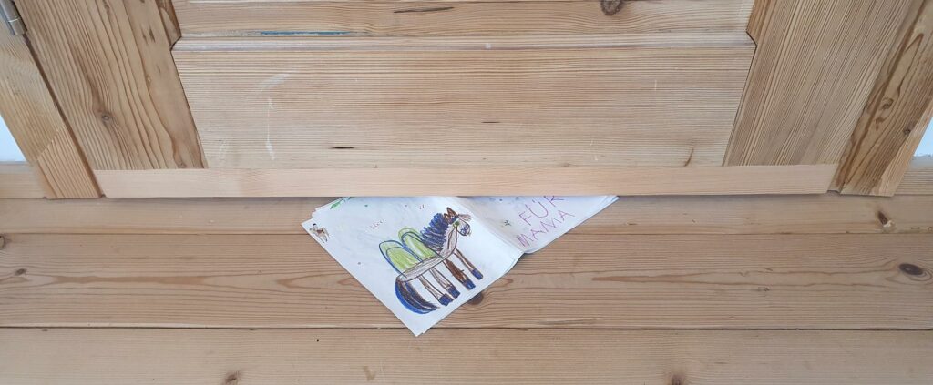 Ein Blatt Papier mit einer Kinderzeichnung (Einhorn) und der Aufschrift "Für Mama" liegt auf einem hellen Holzboden, zur unter einem Türspalt. Die Tür ist ebenfalls aus hellem Holz.