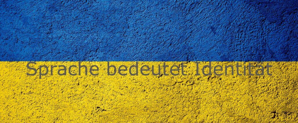 Man sieht die ukrainischen National- bzw. Flaggenfarben Blau und Gelb übereinander auf eine Mauer gemalt. Darüber steht der Spruch "Sprache bedeutet Identität"