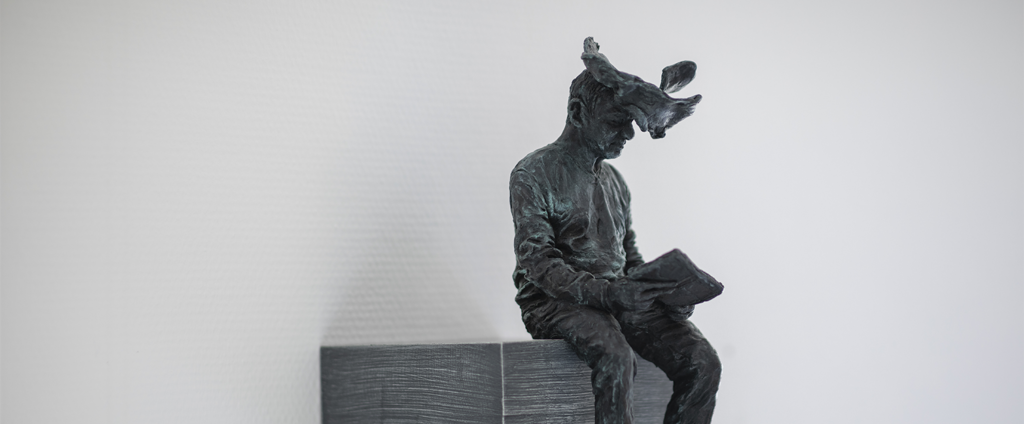 An einer weißem Wand hängt ein grauer Sockel auf dem eine männliche Bronze-Figur sitzt, die in einem Buch liest. Ihr Kopf ist mit einer Wolke umhüllt, die ihre Gedanken symbolisiert.