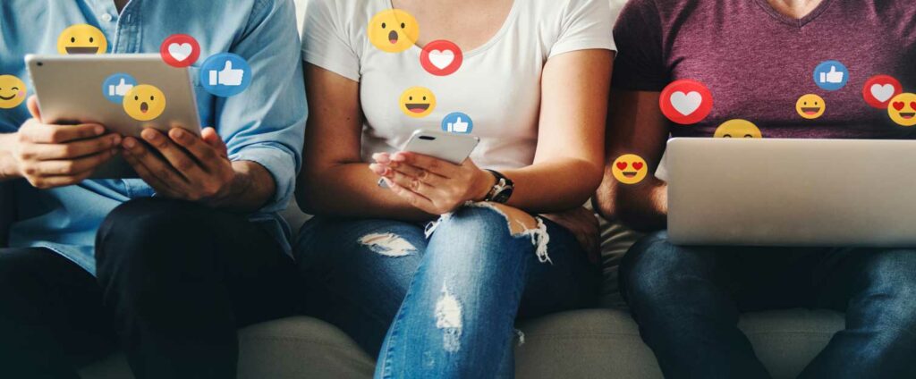 Drei Personen, deren Köpfe nicht zu sehen sind, sitzen auf einer Couch und haben Handys in der Hand. Verschiedene Emojis fliegen drumherum.