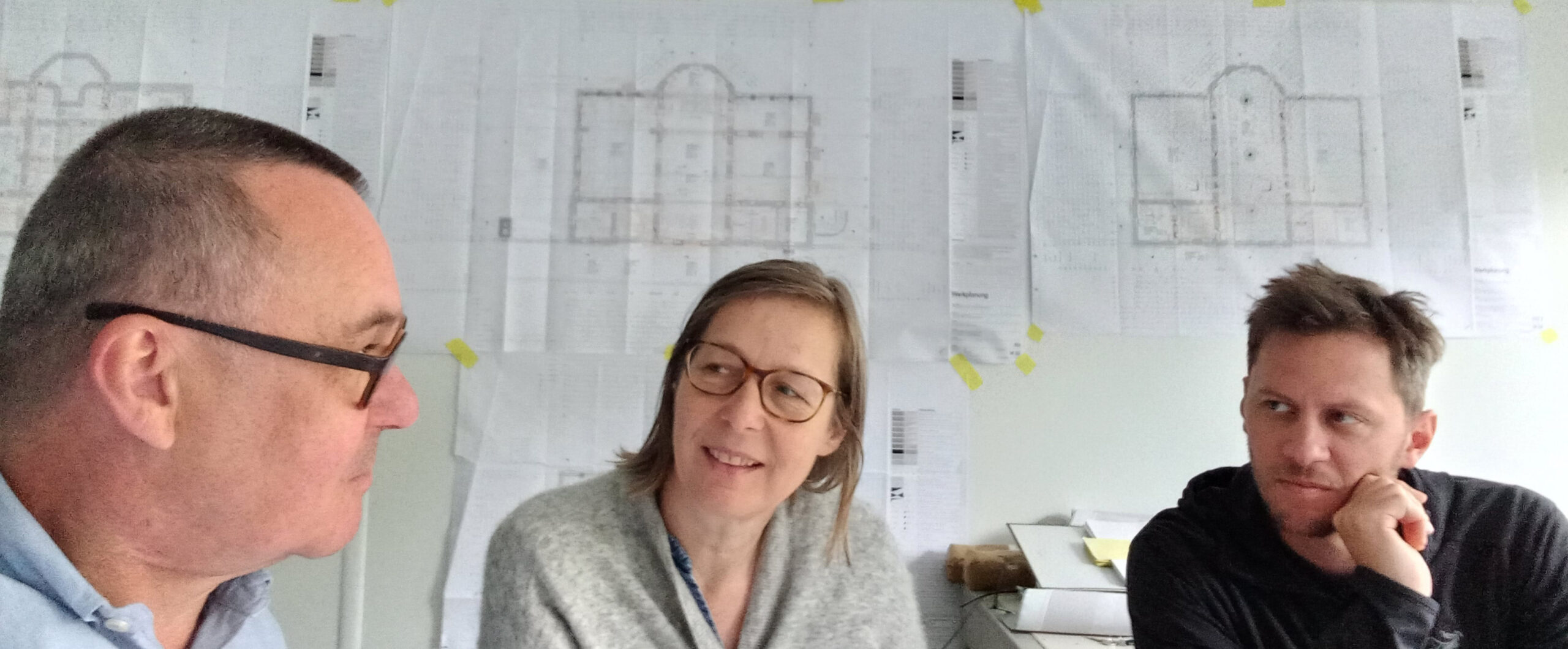 Frank Martin Siefarth vom Medienbüro DiE WORTSTATT sitzt mit dem Team des Alpinen Museums vor einer Wand mit Bauskizzen und erläutert die Ideen für die nächste Sonderausstellung.