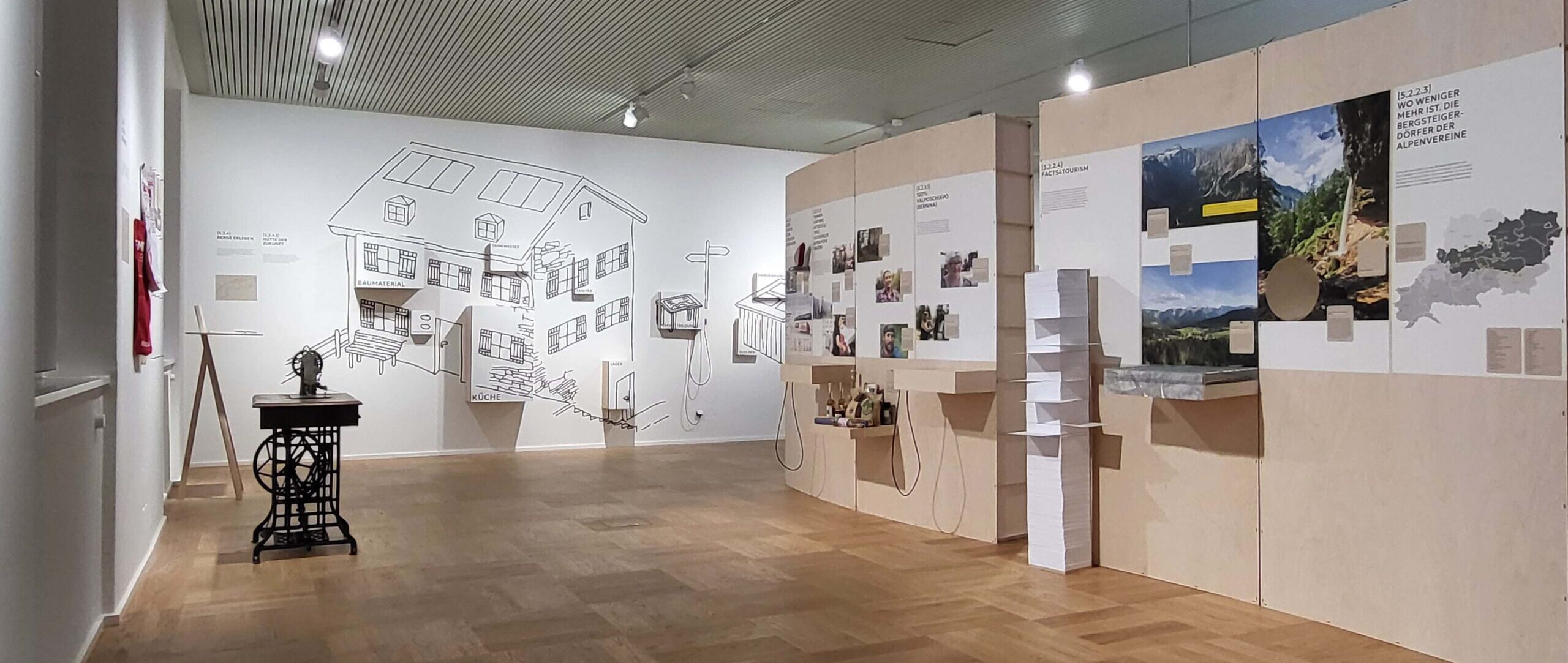 Blick in die Sonderausstellung "Zukunft Alpen"/Kapitel Handeln im Alpinen Museum in München. Zu sehen sind Wände mit vielen Fotos, gegenüber eine historische Nähmaschine und dahinter eine Wand mit einer gezeichneten Berghütte